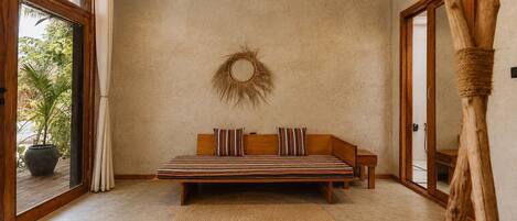 A bedroom with a bed, a round woven wall hanging, a wooden bench, a woven rug, and a large window with a view of greenery.