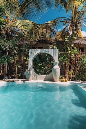 A clear blue pool surrounded by tropical vegetation and a large decorative stone structure. - THE GARTEN LIVING HOTEL (Paje)