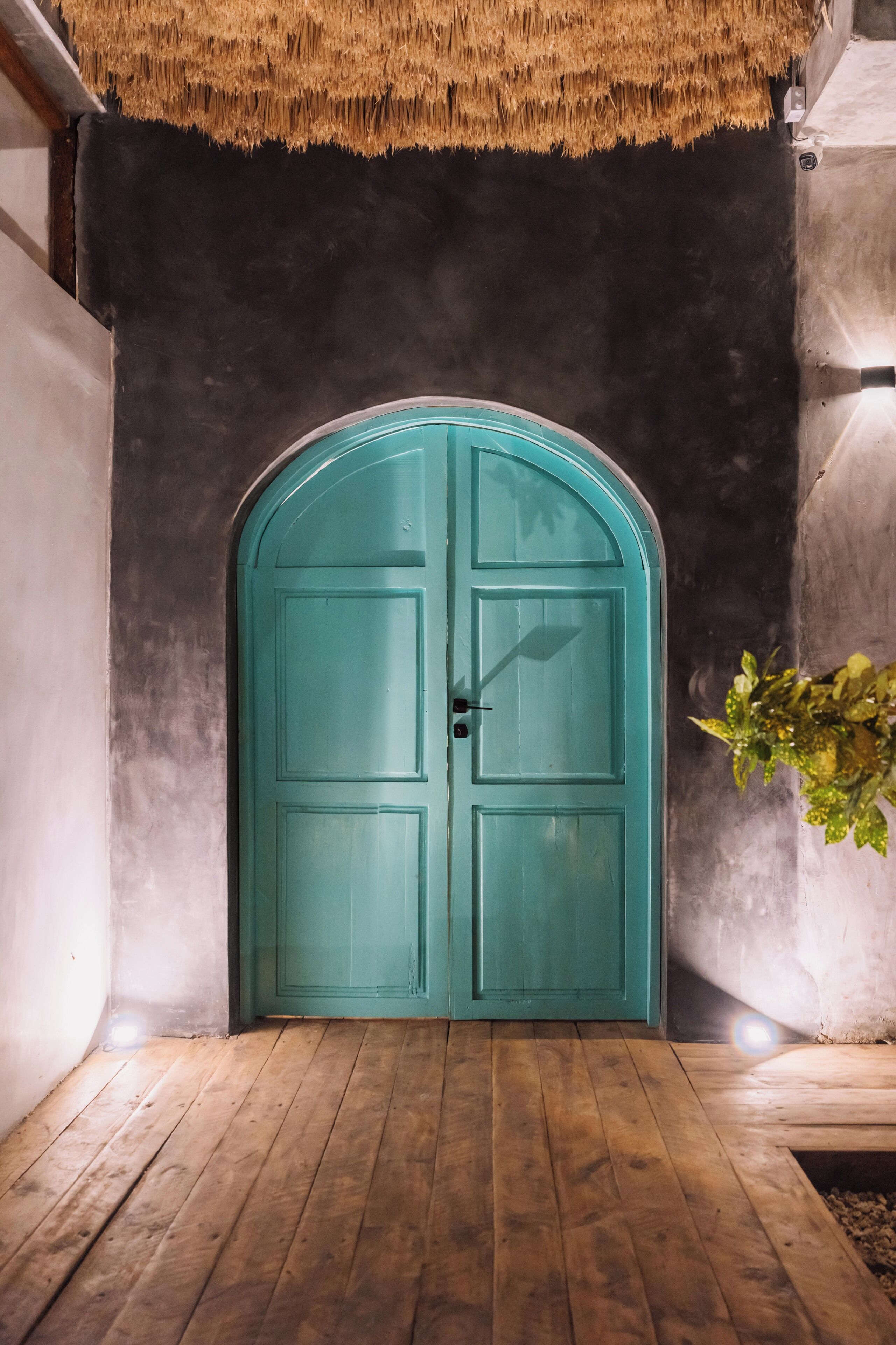 A teal double door with a rounded top, set in a hallway with wooden flooring and a concrete wall.
