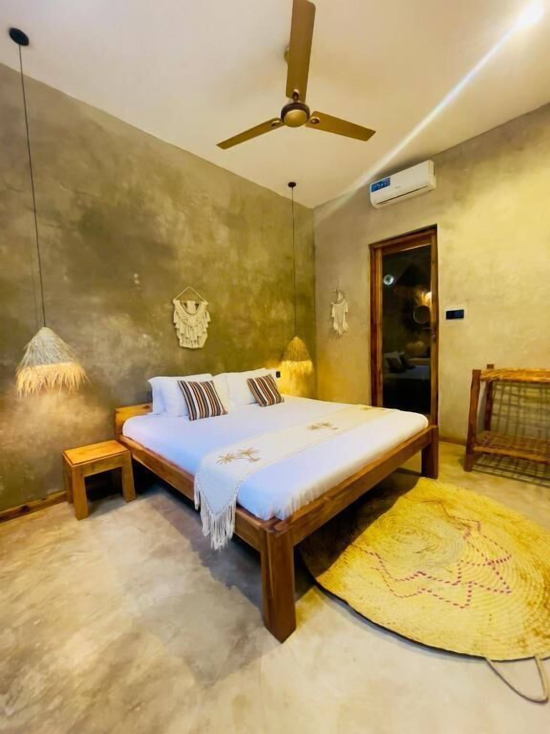 A bedroom with a bed, a ceiling fan, a hanging light, a small wooden nightstand, a woven rug, and a door with a glass panel.