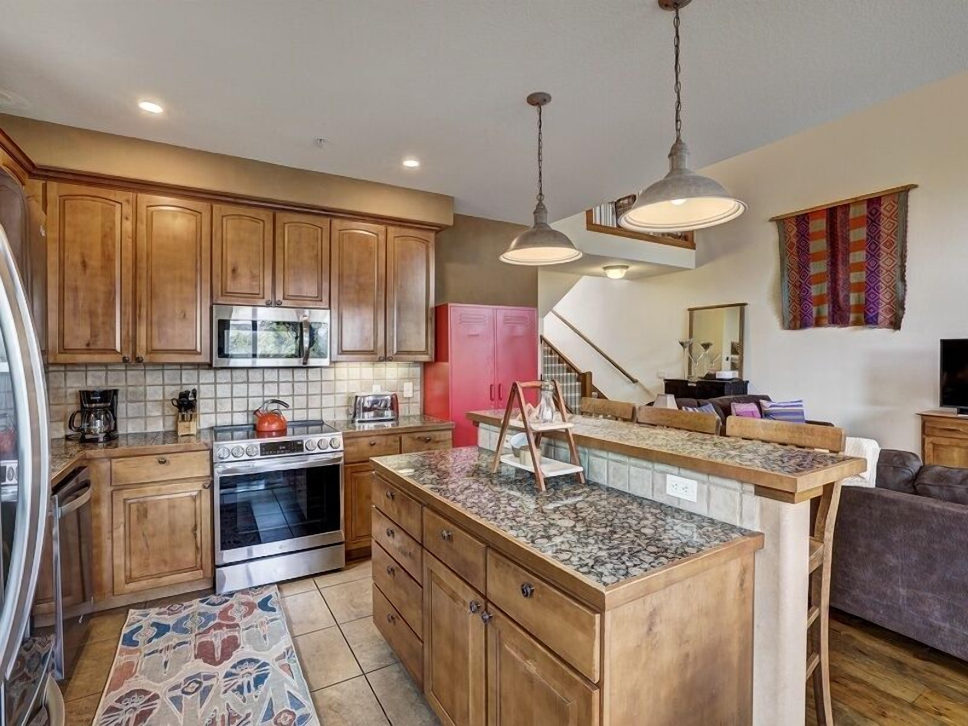 A kitchen with wooden cabinets, a microwave, a stove, and a coffee maker. There is a small island with a sink and a rug on the floor.