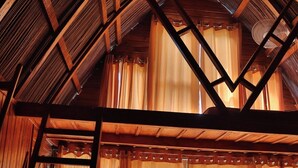 Une chambre mansardée avec des poutres en bois, une échelle et des rideaux légers.