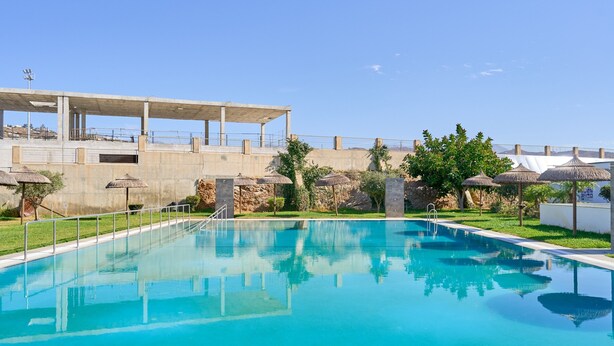 A swimming pool with a concrete deck, surrounded by grass and trees, and a building with a flat roof in the background. - Apartment 'Sea and Mountain Views' with Sea View, Shared Pool, and AC (Torrox)