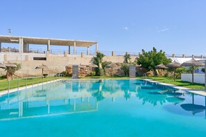 Una piscina con borde de hormigón, rodeada de césped y árboles, y un edificio con tejado plano al fondo.