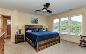 A bedroom with a wooden bed, two bedside tables, a ceiling fan, and a large window with a view of greenery. - Big Canoe 5BR Mountain Escape with Master on the Main – Steps to Amenities (Jasper)