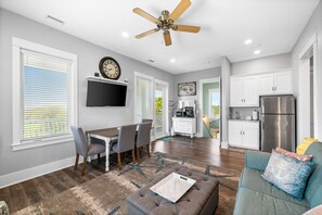 A modern kitchen with a stainless steel refrigerator, a dining area with a table and chairs, a living room with a sofa and ottoman, and a ceiling fan.