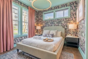 Une chambre à coucher avec un lit, deux tables de chevet, une fenêtre et un mur recouvert d’un papier peint à motifs floraux.