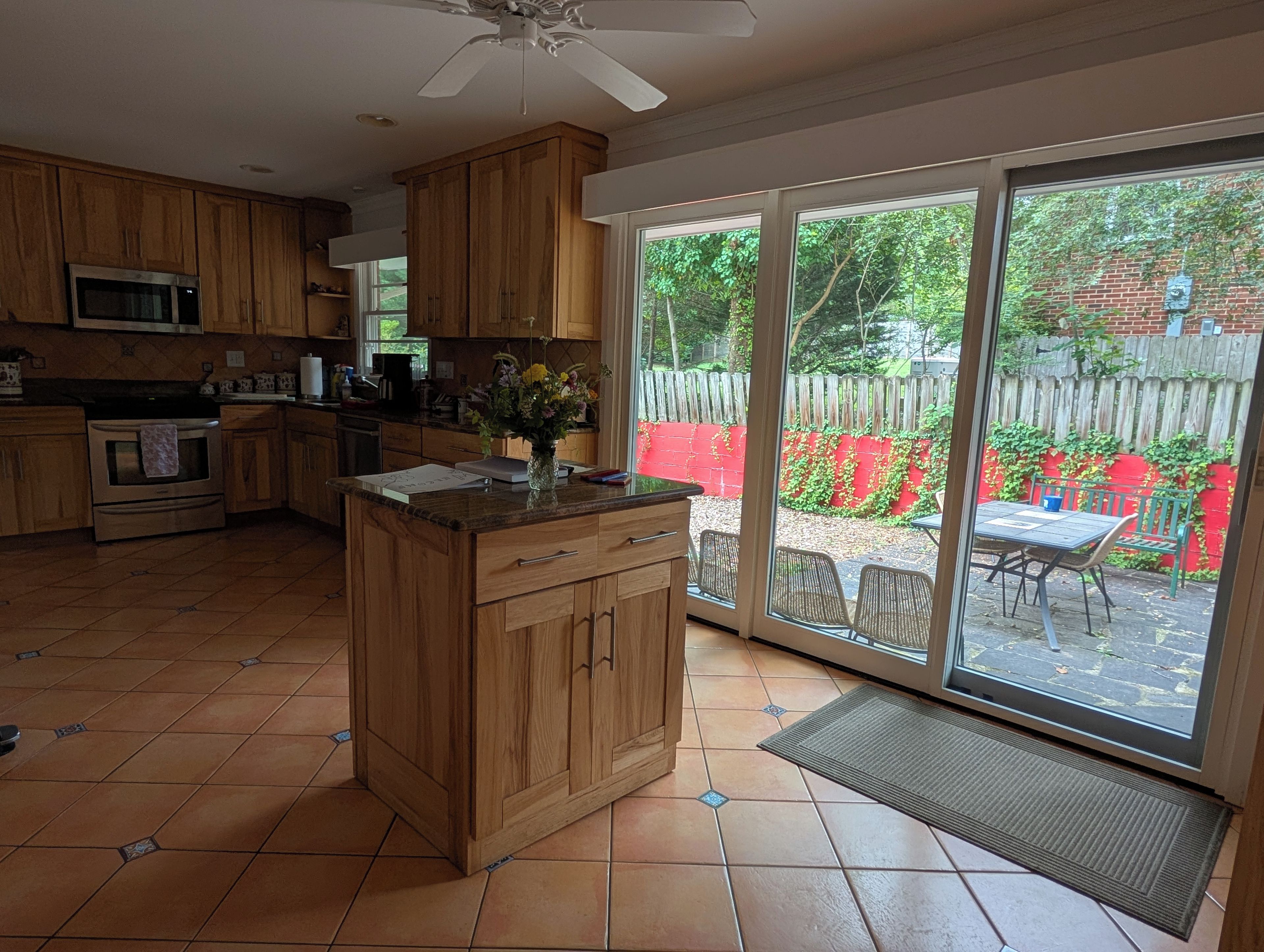 A kitchen with wooden cabinets, a microwave, and a stove. There is a large window with a view of a red fence and greenery outside.