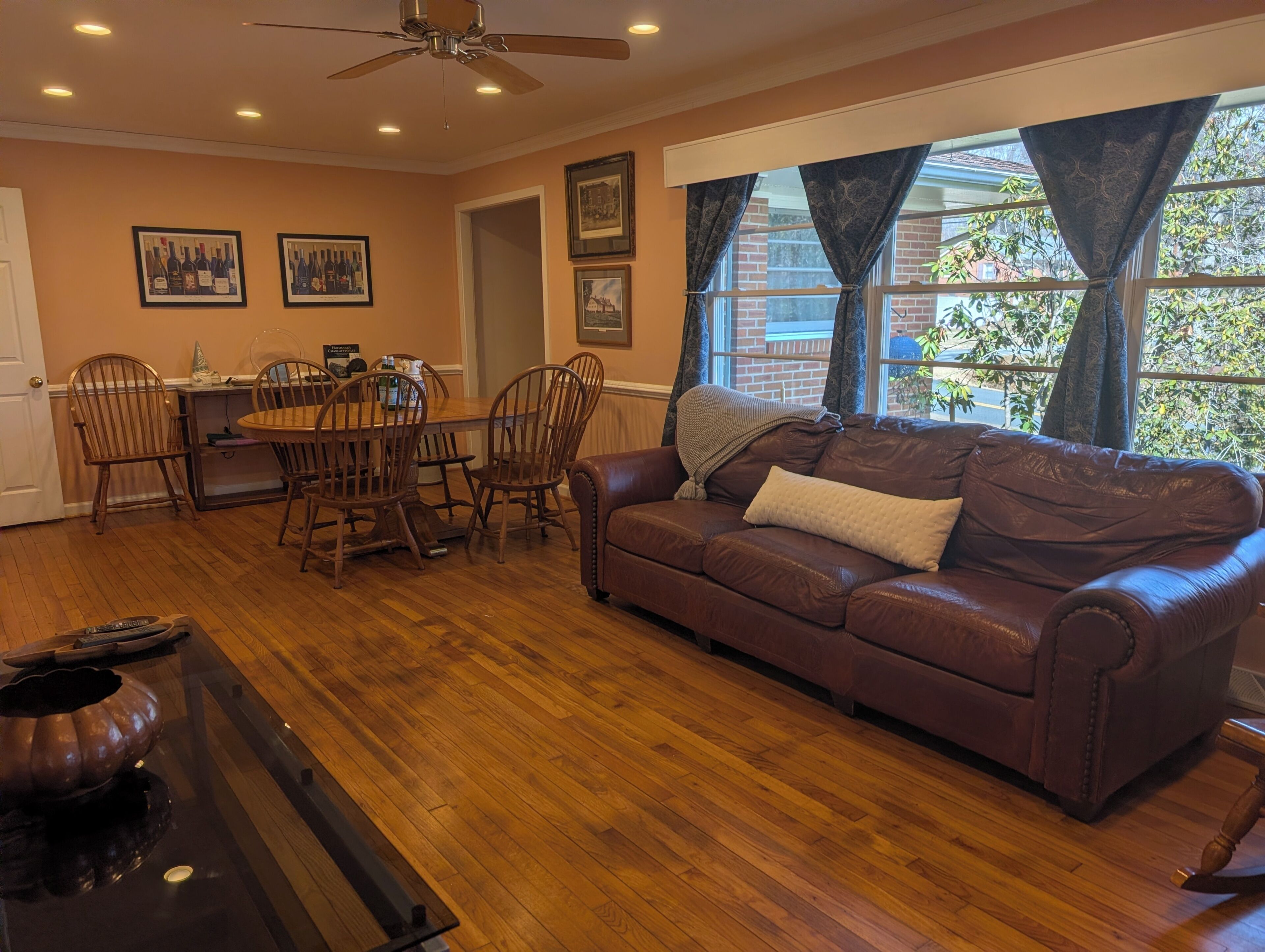 A living room with a brown leather sofa, wooden dining table, and chairs. There is a ceiling fan, framed pictures on the wall, and a window with curtains.