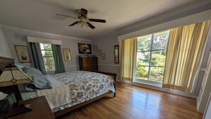 Un dormitorio con suelo de madera, una cama con colcha estampada, un ventilador de techo y un ventanal con cortinas.