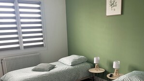 Et soveværelse med to senge, en grøn væg, et vindue med skodder, en radiator og et lille bord med en lampe.