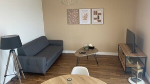 En moderne stue med en grå sofa, et træsofabord, et lille sofabord med et glas og en plante, en lampe, et fjernsyn på et træstel og billedkunst på væggen.