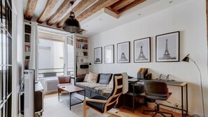 A modern living room with a sofa, a desk, and a bookshelf. There are framed pictures on the wall and a window with a view of the outside. - Athos I Stylish 1BR Home on the Rive Gauche (Paris)
