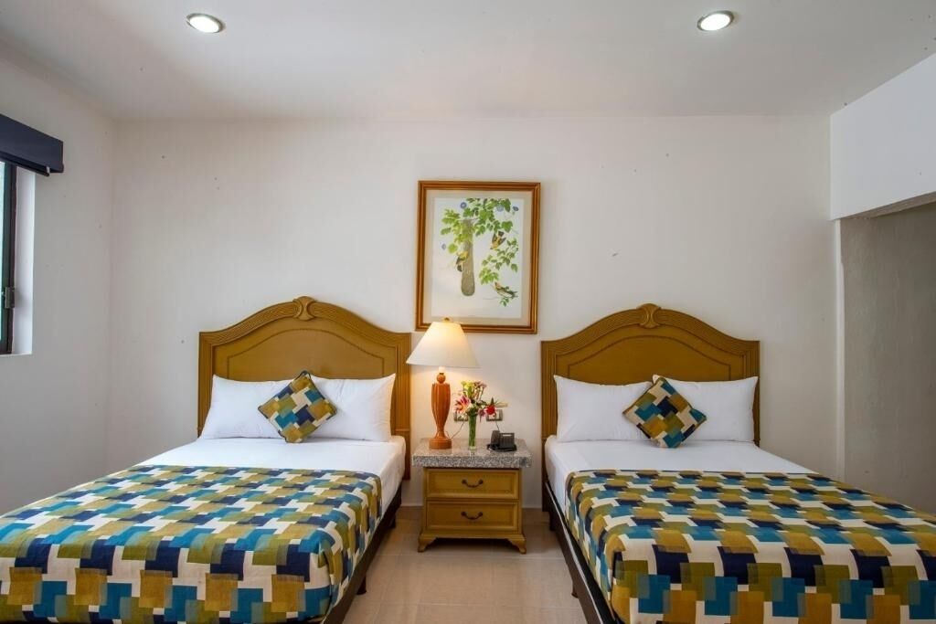 Habitación de hotel con dos camas, cada una con una colcha estampada, una mesita de noche con una lámpara y un jarrón de flores, y un cuadro enmarcado en la pared.