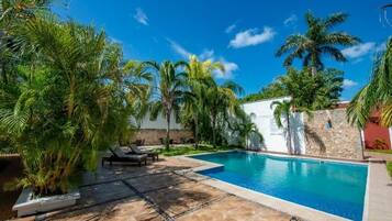 Piscina rodeada por palmeiras e um muro de pedra, com espreguiçadeiras e um terraço com ladrilhos.