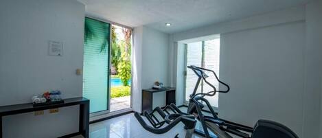 A home gym with a treadmill, elliptical, and weights, a small table with towels, and a window with a view of greenery.