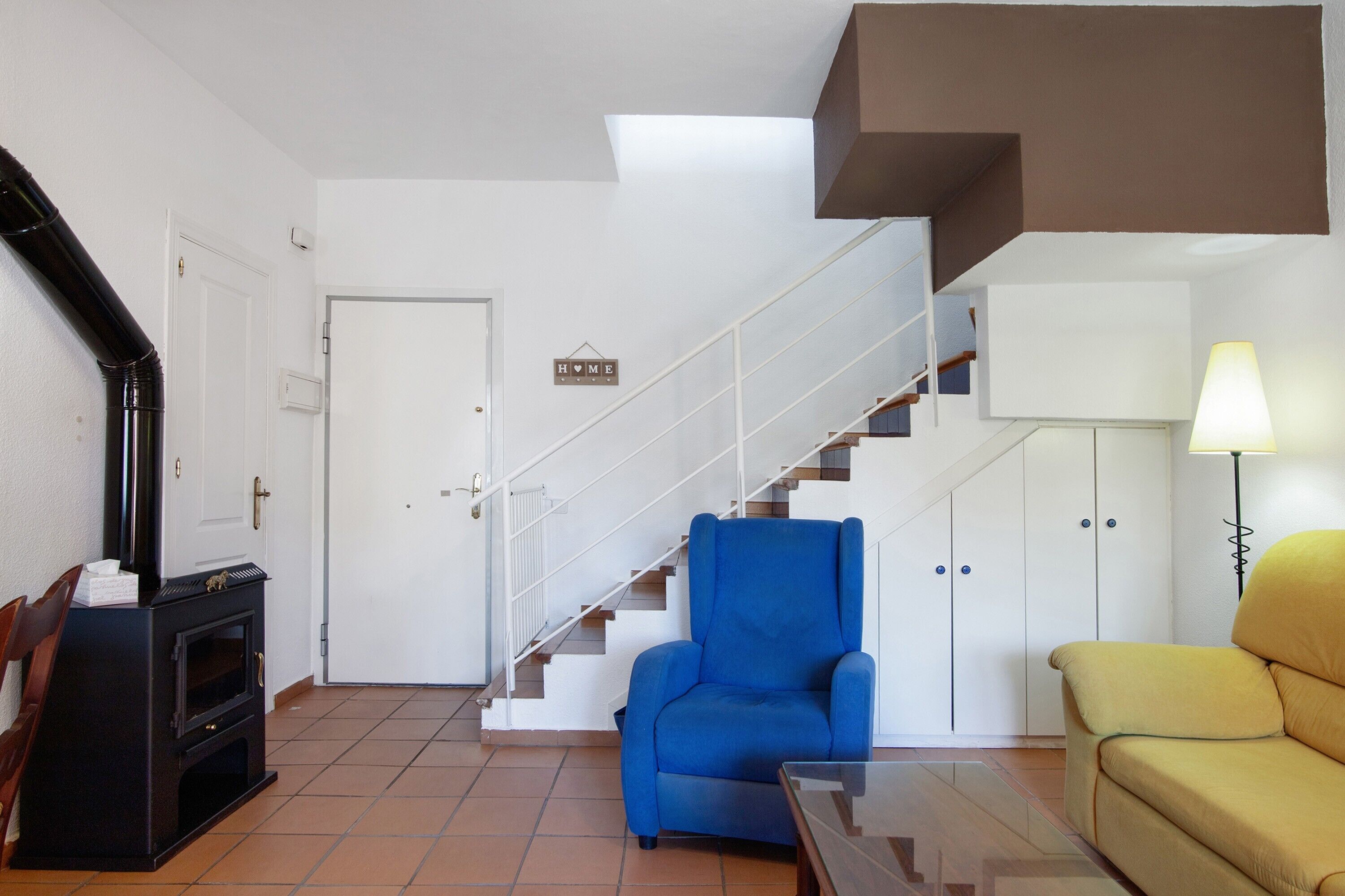 A living room with a blue armchair, a yellow sofa, a fireplace, and a staircase.