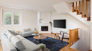 A modern living room with a sofa, coffee table, and fireplace. There is a staircase with wooden railings and a flat-screen TV mounted on the wall.
