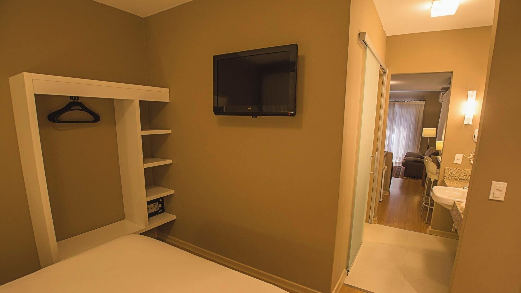 A hotel room with a bed, a wardrobe with shelves, a flat-screen TV, and a bathroom with a sink and mirror.
