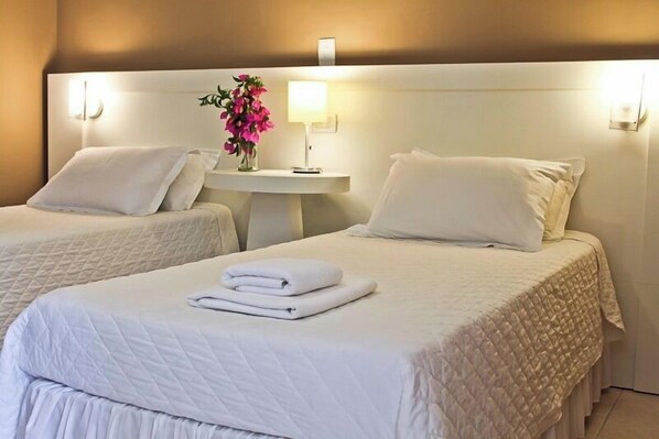 Dos camas ordenadas con sábanas y almohadas blancas, una mesita de noche con lámpara, y un jarrón con flores rosadas.