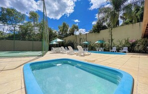 Piscina con sillones de descanso y sombrillas, rodeada por una valla y follaje.