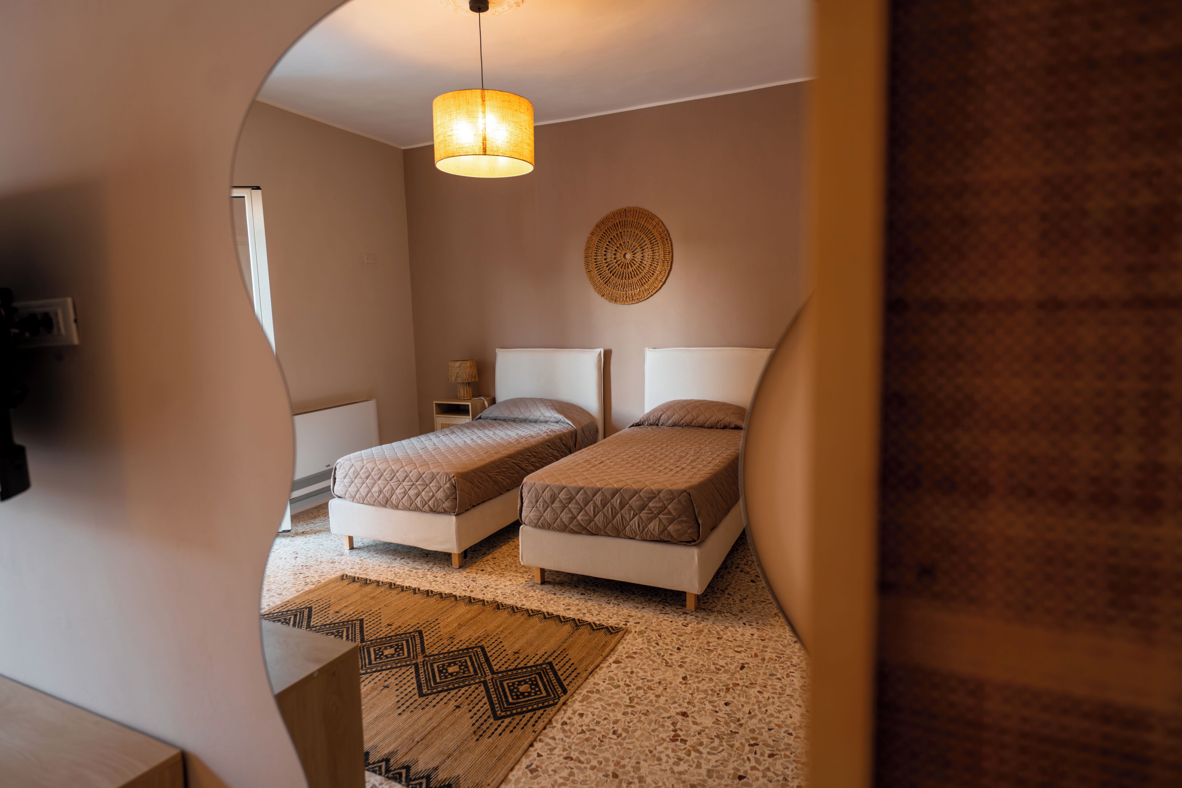 A bedroom with two beds, a hanging light fixture, a woven wall decoration, and a patterned rug.