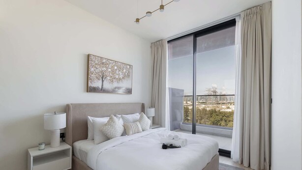A modern bedroom with a large bed, a nightstand, a lamp, a painting, and a view of the city through a large window. - JVC Sleeps 4 With Amazing Views and Large Balcony (Dubai)