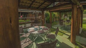 Un patio al aire libre con mesas redondas y sillas de mimbre, un área techada con techo de madera y vista a un jardín.