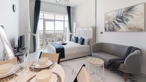 Une chambre d’hôtel moderne avec un grand lit, un canapé gris, un coin repas avec une table dressée pour quatre personnes et une vue sur la ville à travers de grandes fenêtres.