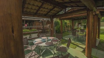 Um pátio externo com mesas redondas e cadeiras de vime, uma área coberta com teto de madeira e vista para um jardim.
