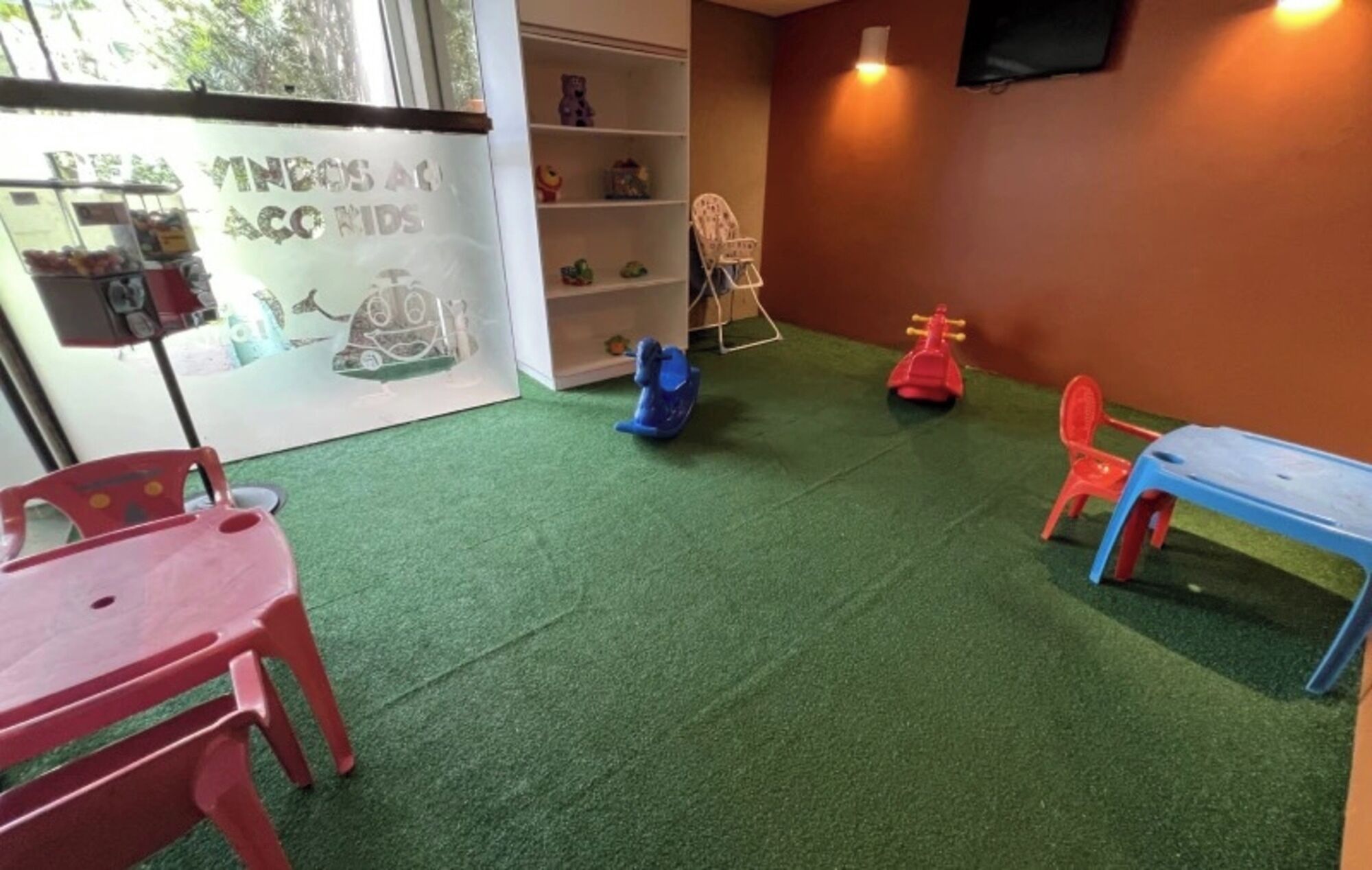 A room with green artificial turf, red and blue plastic chairs, a small table, and a shelf with toys.