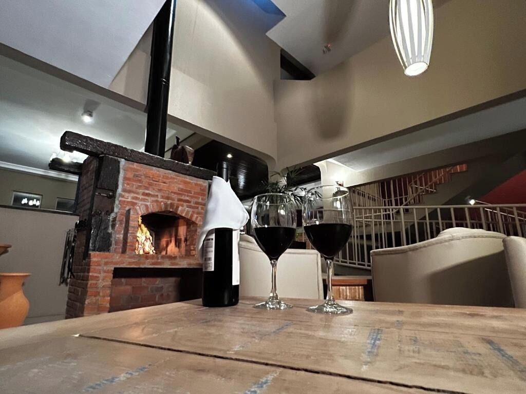 Une table en bois avec deux verres de vin rouge et une bouteille de vin, une cheminée en briques et un escalier en arrière-plan.