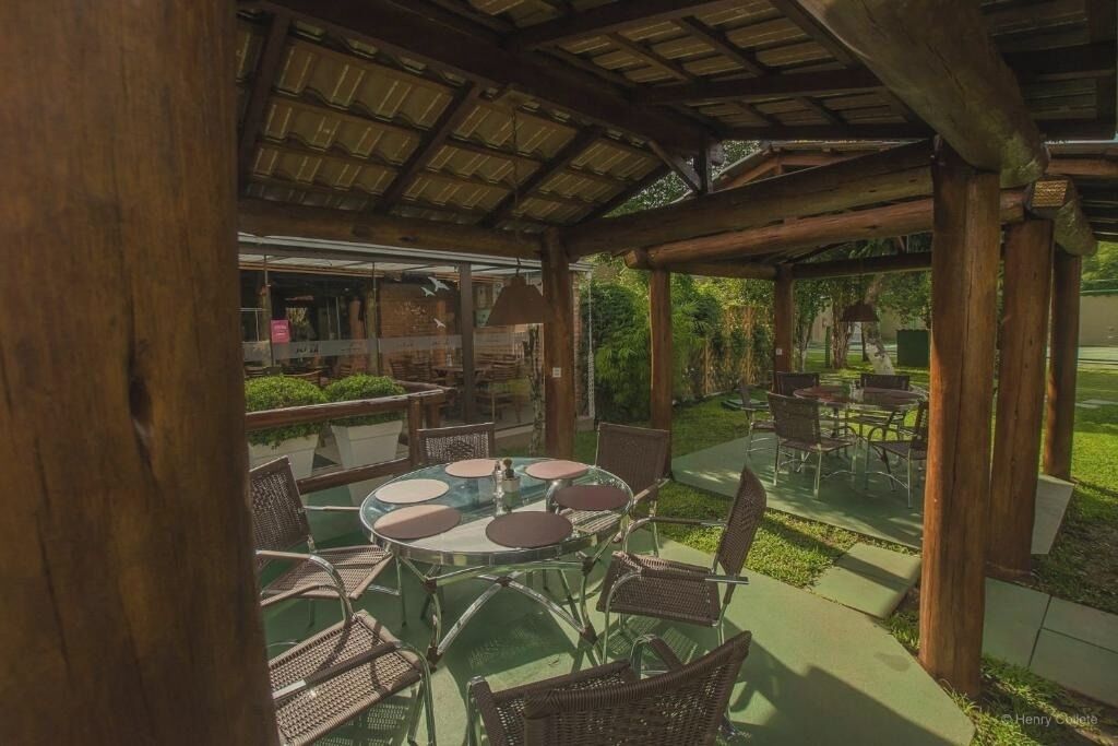 Um pátio externo com mesas redondas e cadeiras de vime, uma área coberta com teto de madeira e vista para um jardim.