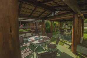 An outdoor patio with round tables and wicker chairs, a covered area with a wooden ceiling, and a view of a garden.