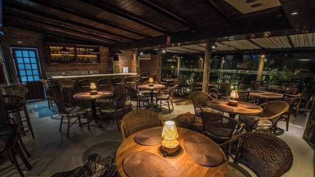 A cozy restaurant with round wooden tables and wicker chairs, featuring a brick wall and a bar area.