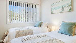 Un dormitorio con dos camas, una ventana con persianas, un cuadro en la pared y una lámpara de noche.