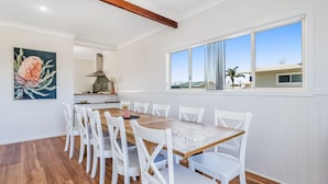 Un área de comedor con una mesa de madera y sillas blancas, un cuadro en la pared y una ventana con vistas a edificios y palmeras.