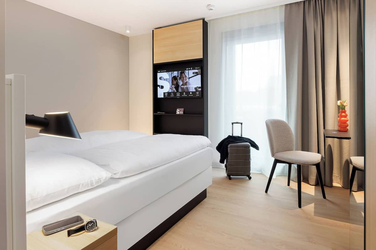 Una habitación de hotel moderna con cama, televisor montado en la pared, una silla, una mesita y una maleta.