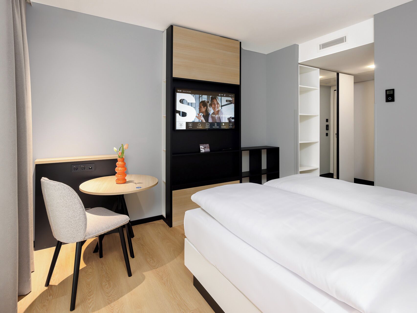 Una habitación de hotel moderna con una cama, un escritorio con una silla, un televisor montado en la pared y una mesita con un florero.