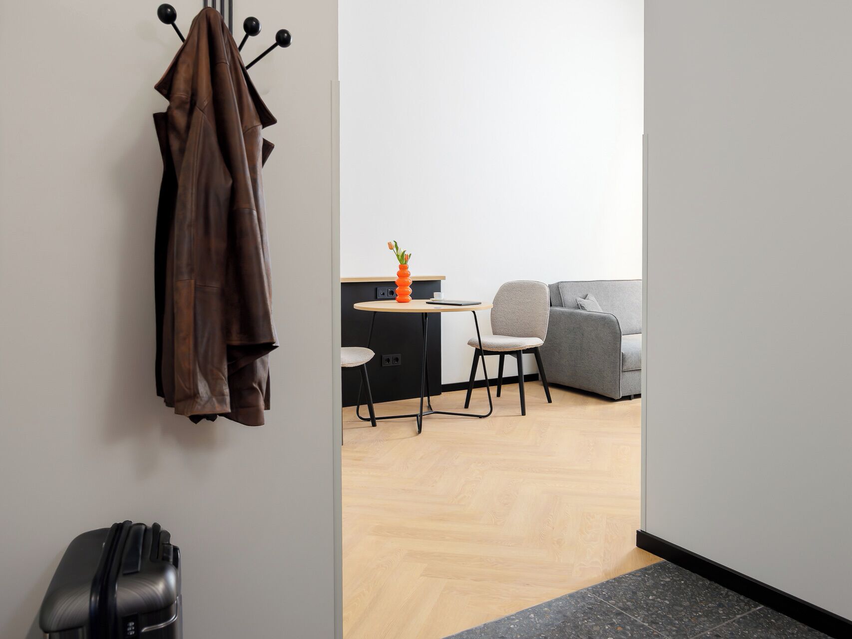 A modern room with a coat rack, a small table with a vase, a grey sofa, and a black suitcase.