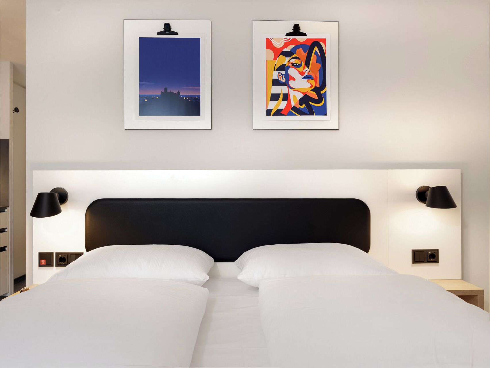 Ein Hotelzimmer mit einem großen Bett, zwei Wandlampen und zwei gerahmten Kunstwerken darüber.