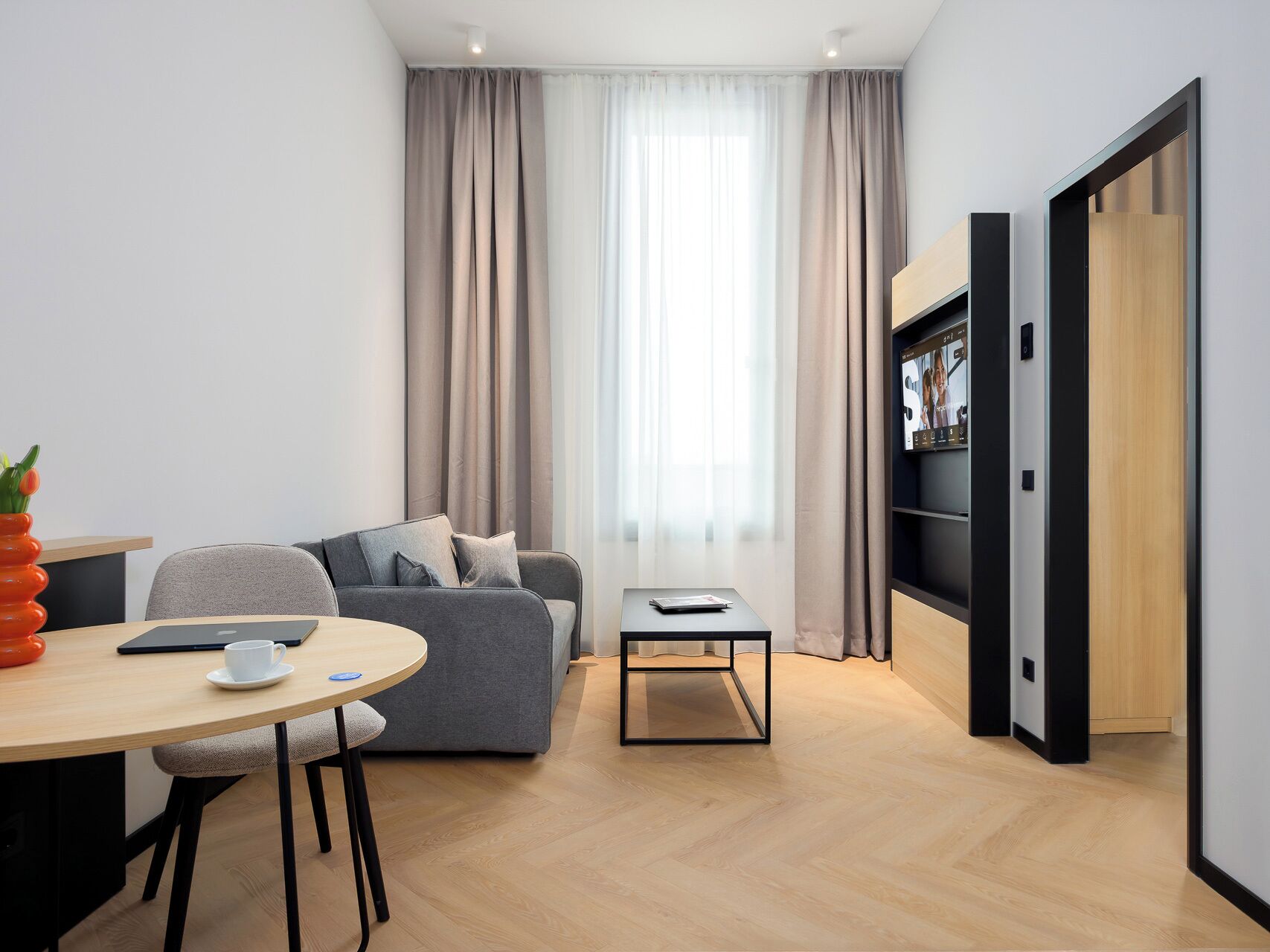 A modern living room with a grey sofa, a small round table with a cup, a bookshelf, and a large window with curtains.