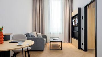 A modern living room with a grey sofa, a small round table with a cup, a bookshelf, and a large window with curtains.