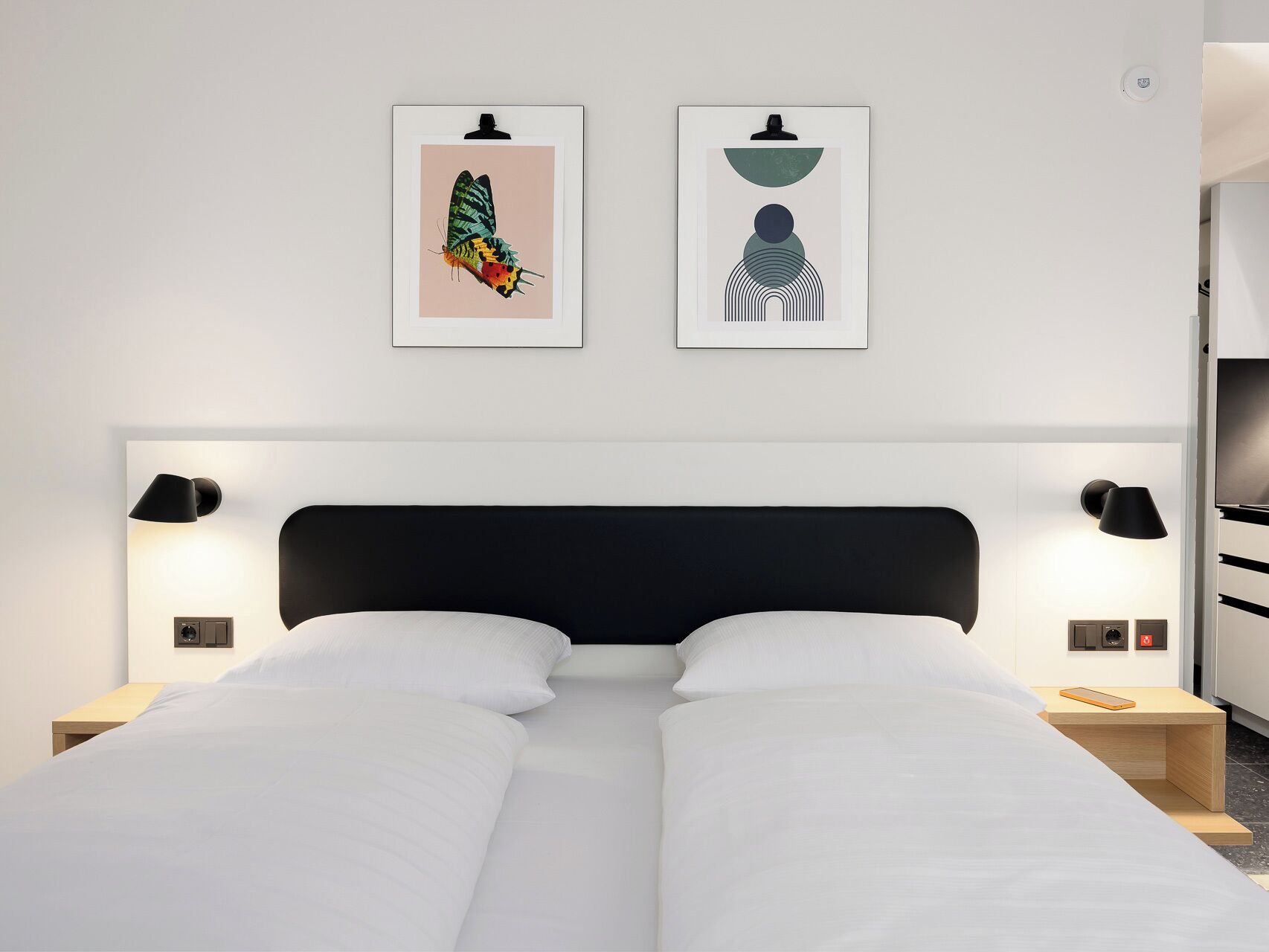 Una habitación de hotel moderna con un cabecero negro, dos mesitas de noche con lámparas, una cama con ropa blanca y dos cuadros enmarcados en la pared.