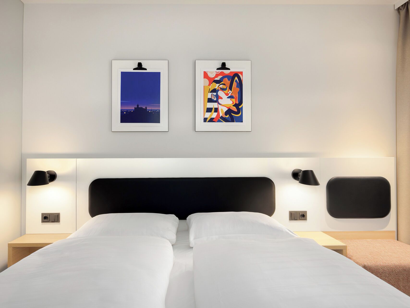 Habitación de hotel con una cama, dos lámparas fijadas a la pared y dos obras de arte enmarcadas.