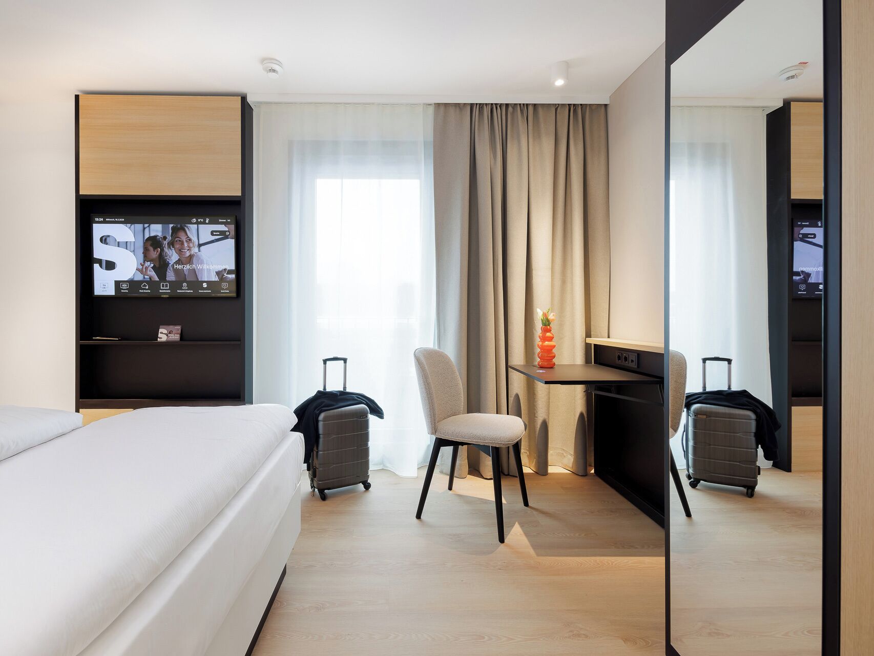 Ein modernes Hotelzimmer mit Bett, Schreibtisch, Stuhl und Fernseher, der eine Willkommensnachricht zeigt.