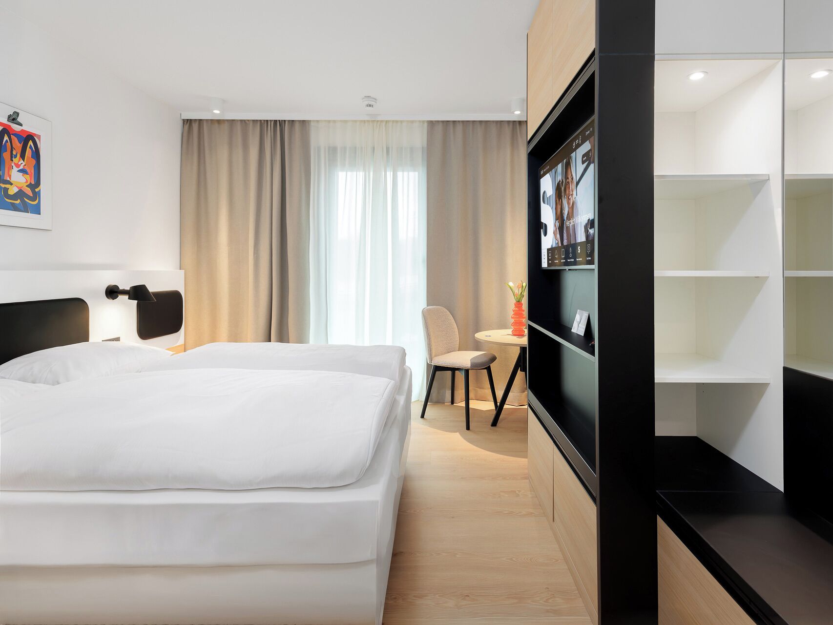 A modern hotel room with a large bed, a desk, a chair, and a TV on the wall.