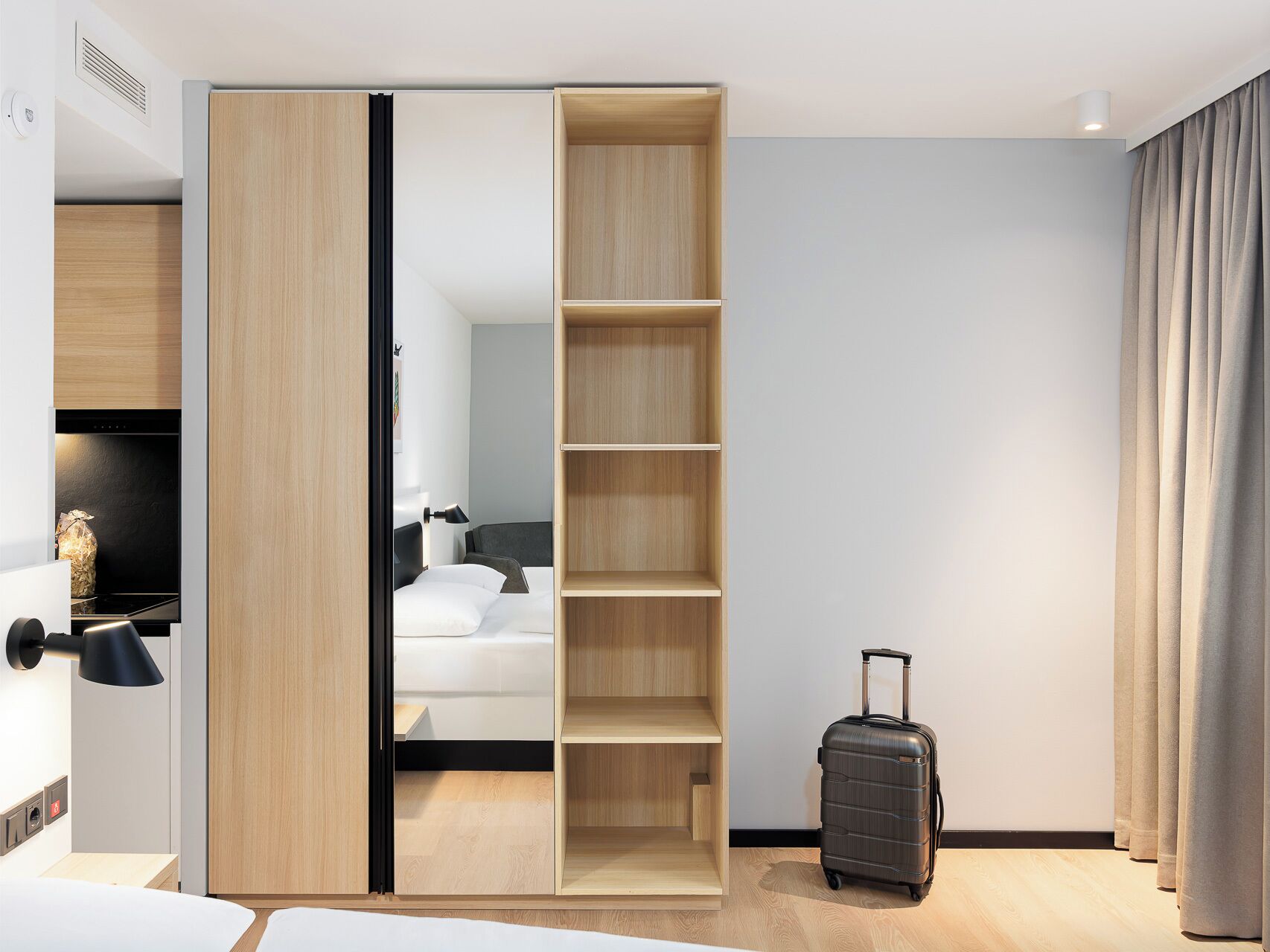 Une chambre d’hôtel avec un placard à portes coulissantes, un lit, un bureau, une valise et une vue sur le couloir.