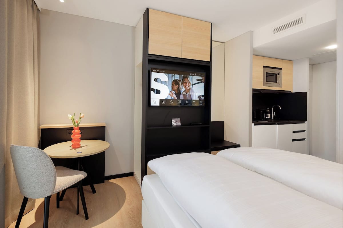 Una habitación de hotel con una cama, un escritorio con una silla, un televisor montado en la pared, un microondas y una pequeña kitchenette.