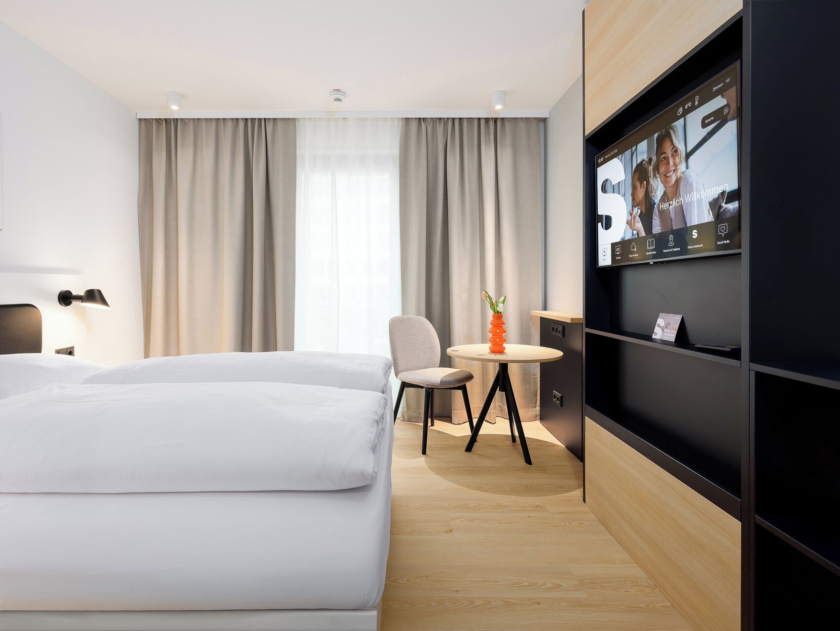 A modern hotel room with a bed, a desk, a chair, a TV, and a window with curtains.
